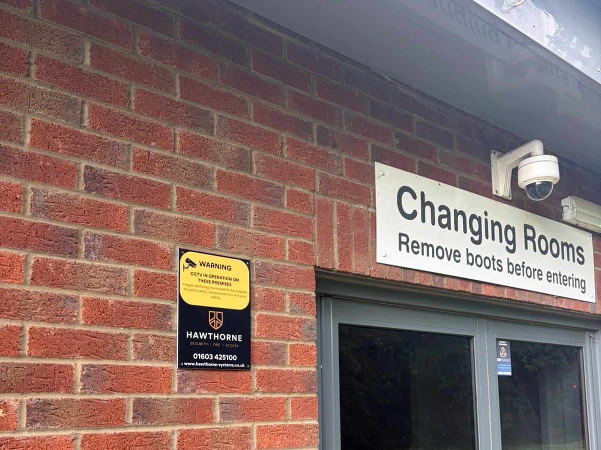 A dome CCTV camera above a sports pavilion changing rooms entrance, with a new Hawthorne CCTV warning sign on the brickwork beside the door.