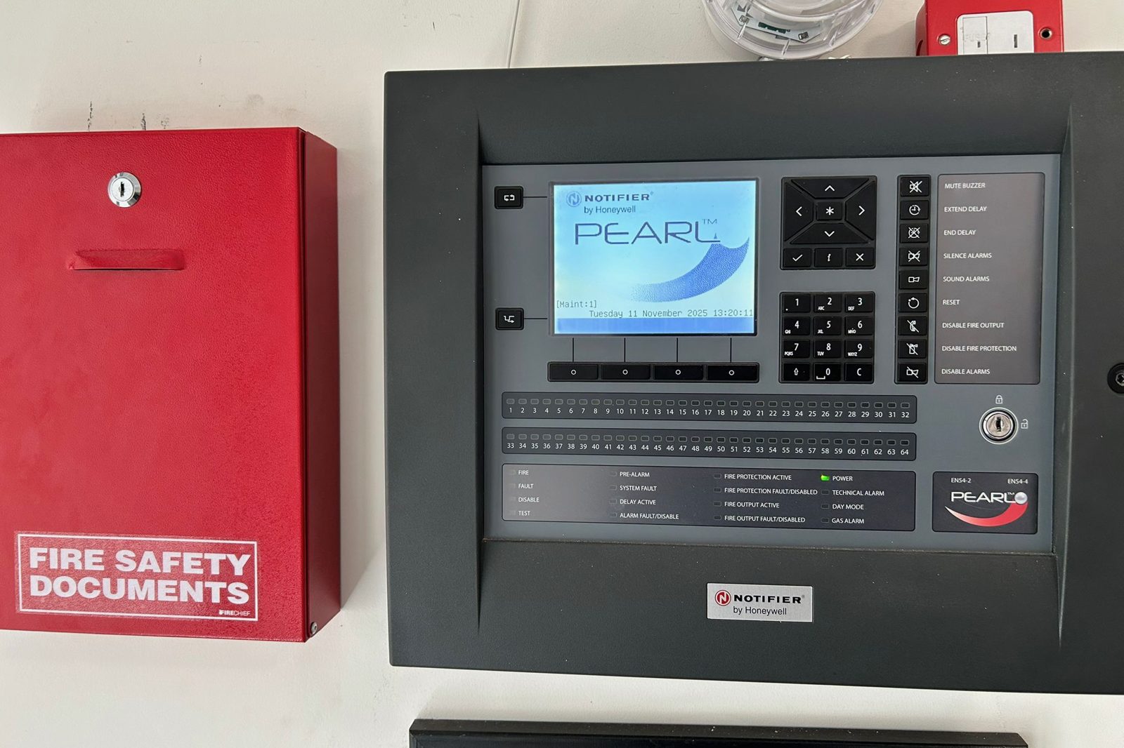 Fire alarm control panel beside a red fire safety documents cabinet, where the log book and certificates are kept.