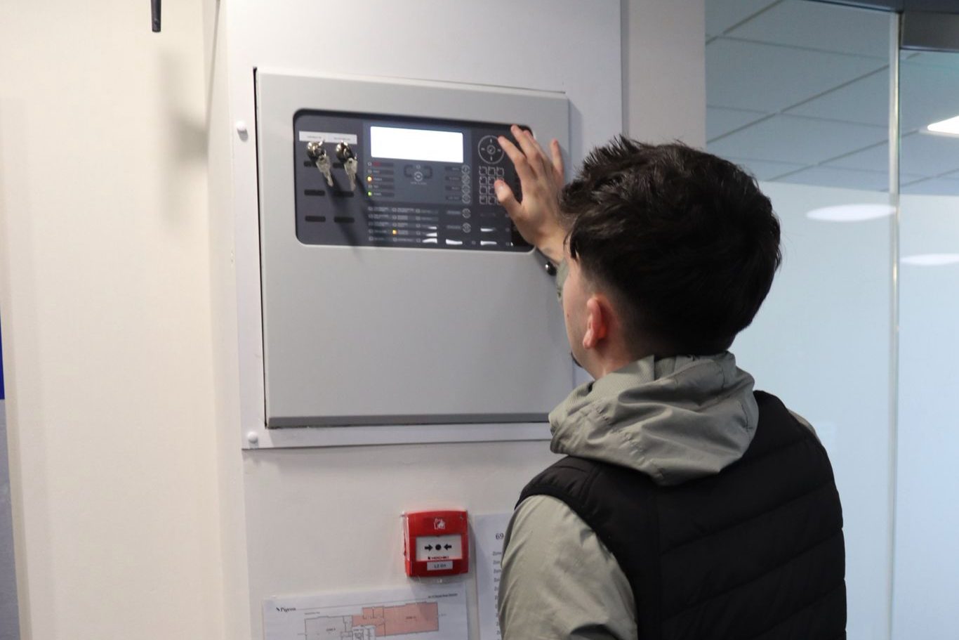 Hawthorne engineer checking a fire alarm control panel during a service visit, with the zone plan and a manual call point below.
