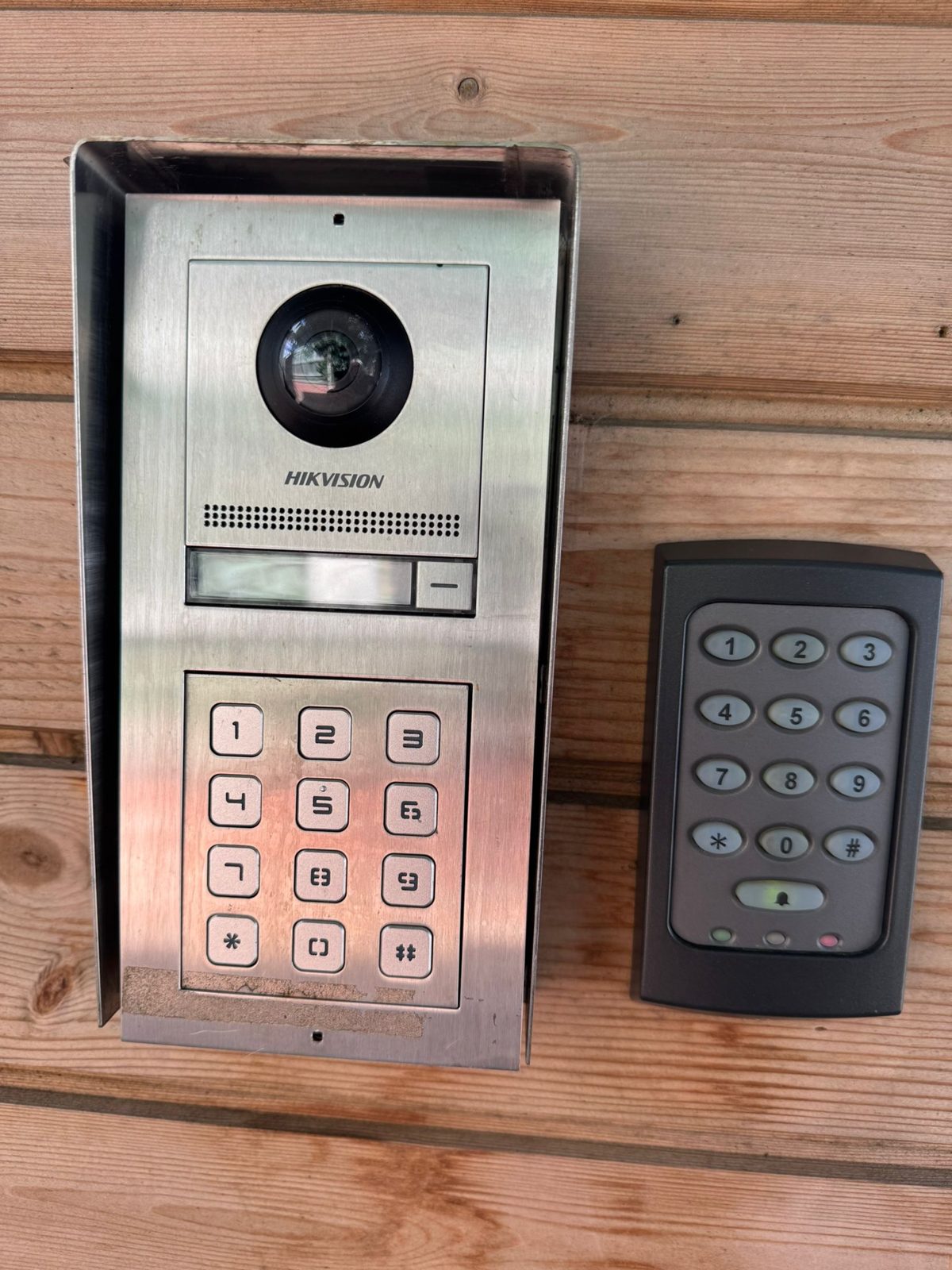 Hikvision video door station beside a keypad reader on timber cladding
